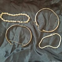 Four silver bracelets displayed on a black fabric background, showing two chain bracelets with lobster clasps and two cuff bracelets, one with a safety chain attachment.