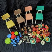 Four plastic Barbie Dream Kitchen chairs in yellow, tan, pink, and teal with a large assortment of miniature kitchen accessories including colorful plates, cups, saucepans, utensils, and plastic food pieces arranged in front.