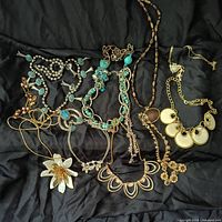 Photo showing 11 bold statement necklaces laid out on black fabric, including imitation turquoise squash blossom style necklace, large white flower pendant necklace, gold-tone chain necklaces with floral and coin pendants, and a sparkly fox pendant necklace.