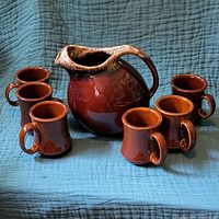Set of one Hull pottery pitcher and six brown mugs displayed on blue cloth