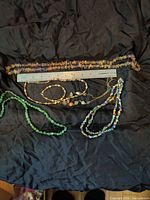 Photo showing all necklaces and bracelet laid on black cloth with a ruler for size reference, depicting a variety of stones in different colors including green, yellow cat's eye, multi-color chip stones, and beige beads.