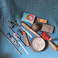 Wide view of all vintage tools laid on blue quilt, showing pocket hand warmer with original box, folding ruler, brass hygrometer, hand drill, screw plates, oil can, and metal box.