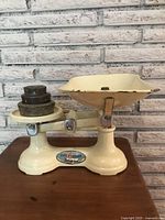 Full view of cream enamel vintage kitchen scale with cast iron weights stacked on left platform, placed on wooden surface and brick background