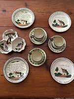View showing three tea cups with saucers, three decorated plates with hand-painted villages and text, and a tri-section divided dish, all ceramic with hand-painted decoration.