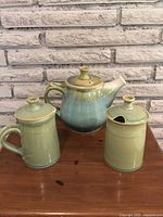 Full view of the three-piece artisan pottery tea set showing the teapot, creamer, and sugar bowl with lids against a brick background.