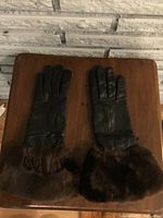 Pair of dark brown women's leather gloves laid flat on a wooden surface with a light brick wall background. Gloves have fur trim at the wrist and show minor wear.