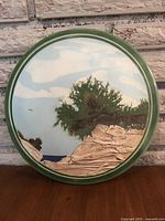 Front view of round ceramic plaque showing hand-painted landscape with trees, rocks, sky, and green border.