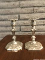 Pair of antique silver plated candlesticks on a wooden surface against a stone wall background, showing detailed patterns and shape.