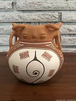 Front view of ceramic pottery vase showing round body with painted geometric designs and sculpted handle figure detail on top.