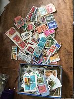 Top view of multiple used postage stamps spread on wooden surface and in a small blue box, showing different colors and designs.