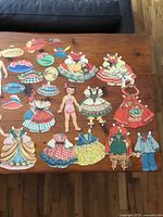 Two sets of vintage cardboard paper dolls with various outfits and hats laid out on wooden table.