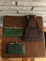 Three vintage leather accessories arranged on wooden surface: two green wallets and one brown trapezoid handbag with brass-tone metal details.