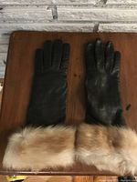 Front view of pair of black leather gloves with light brown fur cuffs placed flat on wooden surface against white brick background.