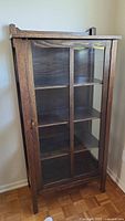 Front view of the antique cabinet showing wood frame and glass door with four shelves inside.