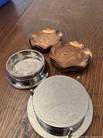 Top view of the silver-plated wine coaster with detailed engravings and a set of copper coasters with leaf engravings stacked on a wood surface.
