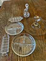 Full view of six vintage crystal items including cake plate with handle, cruet with stopper, two round serving plates, and two rectangular butter dishes with ribbed and spiral cut designs.