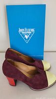 Side view of left and right John Fluevog dress shoes next to John Fluevog box showing burgundy suede upper, yellow toe caps, and red heels.