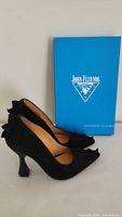 Side view of both black suede John Fluevog Neptune high heels beside blue branded box