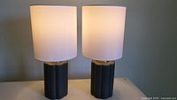Pair of small table lamps on a flat surface, illuminated with white linen-type shades and black ceramic style bases.
