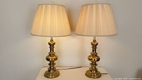 Pair of traditional brass finish metal table lamps with cream drum shades on a flat surface against wall.