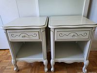Front view showing two white wood nightstands with gold trim, single drawer with metal handle, open lower shelf, curved legs and apron base.