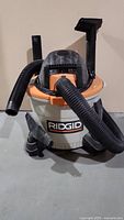 Front view of the Ridgid wet/dry shop vacuum showing the orange lid, white body, flexible black hose connected at front, and Ridgid brand label.