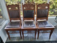 Three solid oak antique chairs in a row with ornate carved backs and original dark leather seats, showing wear and peeling on the seats.
