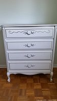 Front view of the white wood dresser with four drawers, ornate metal handles, and gold trim detailing on drawer fronts.