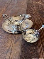 Set of three vintage silver-plated brass serveware items on wooden surface including gravy boat with underplate, small bowl, and spoon.
