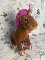 Front and side view of a brown plush unicorn with vibrant pink mane and tail, gold hooves and horn, seated position.