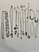 Photo showing all 14 necklaces and 3 pairs of earrings laid out on a surface, showcasing various chain styles and pendant types.
