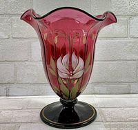 Front view showing the hand-painted white day lily floral design on ruby cranberry glass with ruffled edge and black foot with gold trim