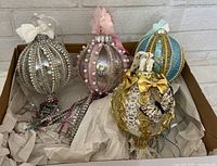 Four handmade whimsical ornaments in a box showing silver, pink, teal blue, and white designs with decorative elements