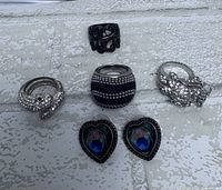 Photo showing a collection of six rings and one pair of heart-shaped peacock design earrings. Rings include rhinestone-encrusted serpent ring, black and silver dotted ring, silver dragon ring, and others.