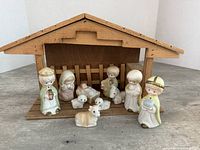 Photo of assembled nativity scene with 6 ceramic figurines and 3 lambs arranged in front of a wooden stable.