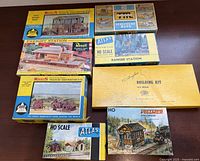 Photo of eight boxed HO scale train model kits arranged on a table showing various brands and building types.