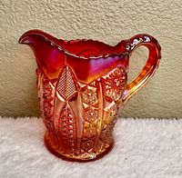 Front-facing view of vintage amberina carnival glass creamer pitcher emphasizing its rich red to amber gradient, iridescent sheen, scalloped rim and handle.