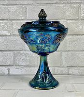 Front view of blue carnival glass compote dish with lid, showing grape and leaf pattern, footed base, and lid knob.