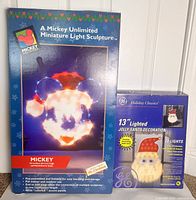 Photo showing the boxed Mr Christmas Mickey Mouse Unlimited light sculpture and boxed GE Holiday Classics Jolly Santa lighted decoration, side by side for scale and packaging condition.