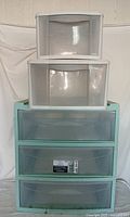 Five plastic storage cabinets stacked to show size and condition, including a larger green cabinet with three drawers and smaller white cabinets with one drawer each.