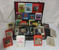 Overview of the 28 assorted 8-track tapes spread out in front of open black plaid carrying case with red lining.