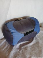 Blue-grey sleeping bag compressed and secured with black straps, lying on a light-colored surface.