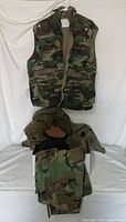 Camouflage vest with multiple pockets and zipper plus snap closure, folded camouflage pants below.