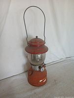 Full view of the red vintage camping lantern standing approximately 12 inches tall against a white backdrop, showing metal handle and overall condition including paint wear.