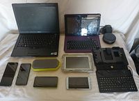 Overview of the electronic devices arranged on a white surface including laptop, tablet with keyboard, multiple smartphones, Bluetooth speaker, wireless keyboard, charging dock, and smart speaker.