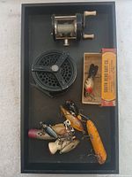 One photo showing two fishing reels, one conventional metal reel with wooden handles and one black fly reel, along with eight assorted fishing lures including one boxed lure.