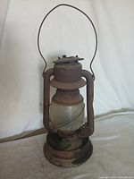 Full view of the rusty metal hurricane lantern with handle and cracked globe.