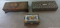 Photo showing top view of three jewelry boxes: gold-tone rectangular, silver-tone rectangular with floral embossing, and small decorative box with floral porcelain lid.
