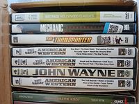 Photo showing DVD spines stacked in a box including AMC Hollywood Classics Rat Pack 4 movies on 2 DVDs, Mechanic Resurrection, The Transporter, and multiple John Wayne Western movie collections.