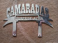 Front view of chrome metal license plate accessory with 'CAMARADAS SURPRISE AZ' text and mounting tabs